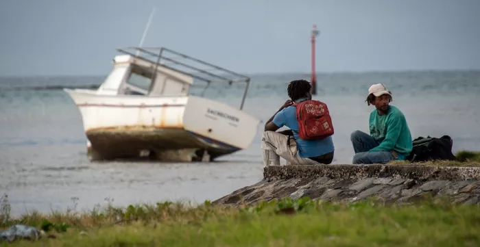 Two young fishers sit by the sea in front of their boat on the sand.