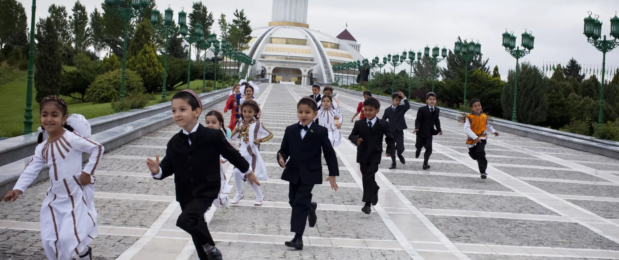 Young children in traditional outfits run in front of the Independence Monument of Turkmenistan in Ashgabat, Turkmenistan.