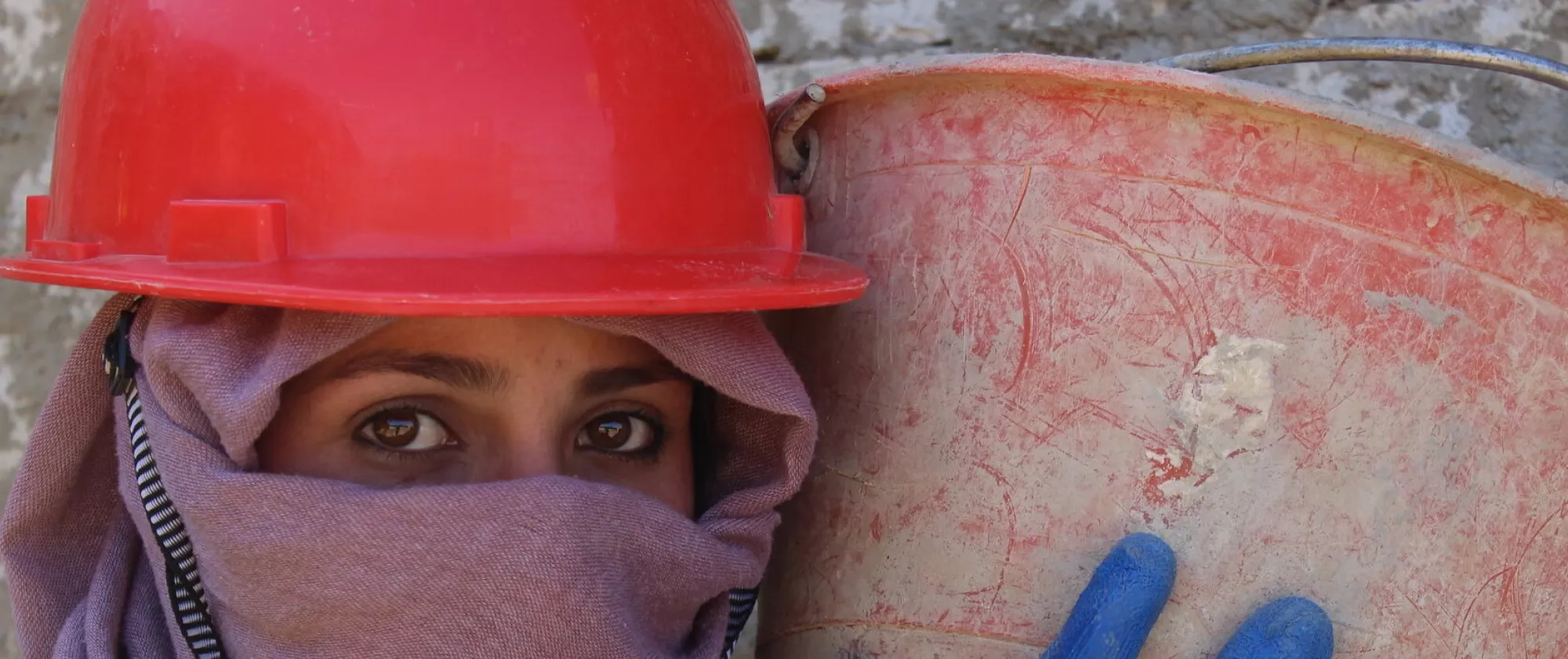 A woman with a red hard hat, holding bucket of concrete on her shoulder at a construction site