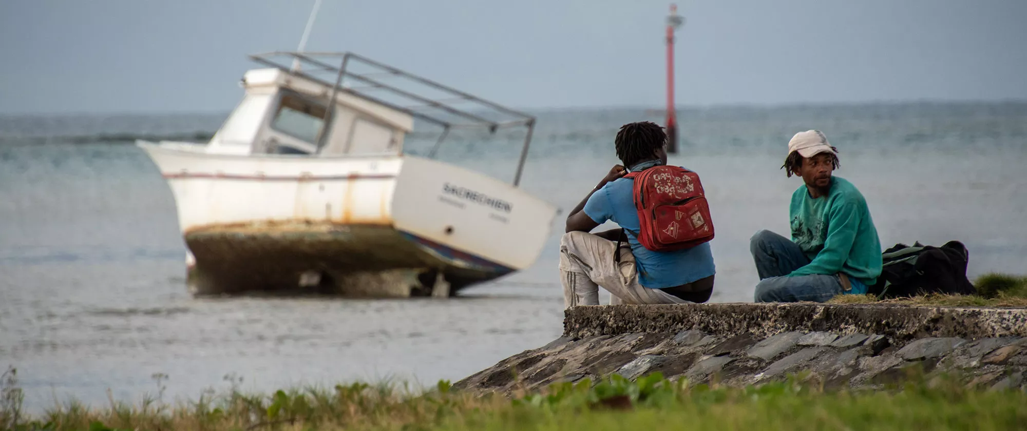 Two young fishers sit by the sea in front of their boat on the sand.